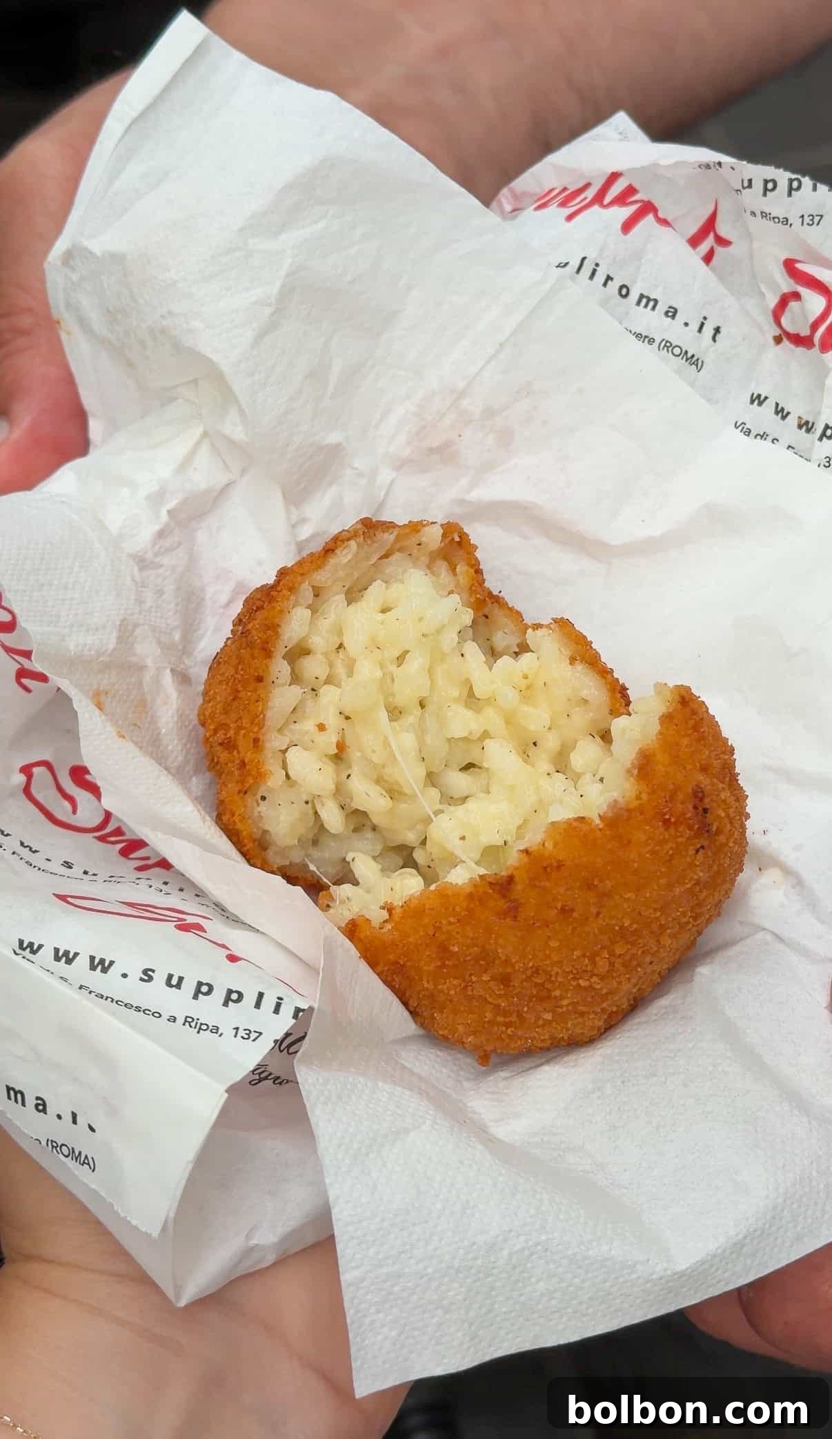 A hand holds a half-eaten, golden-brown fried rice ball with a creamy rice filling, wrapped in a white napkin. The rice ball is resting on a branded paper with red and black text.