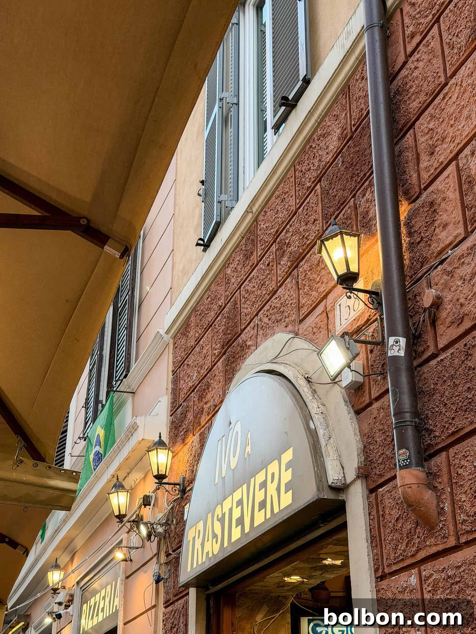 A street view of a building in Rome with a sign reading "IVO A TRASTEVERE" above an arched entrance, illuminated by wall lanterns, and an adjacent "PIZZERIA" sign visible to the left.
