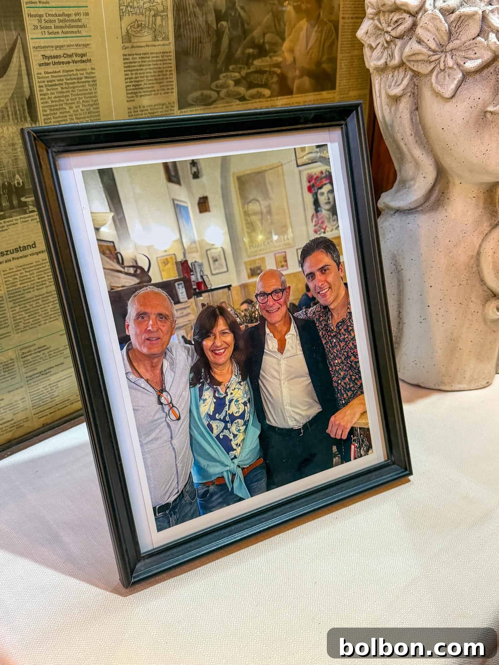 A framed photo of four smiling adults standing close together indoors, with their arms around each other, displayed on a table next to a decorative vase and in front of background newspaper clippings.