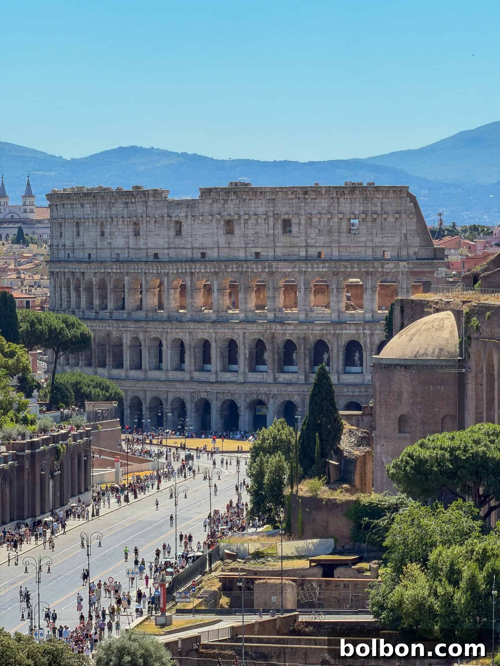 The Colosseum in Rome stands under a clear blue sky, surrounded by crowds of tourists and nearby historic ruins, with distant mountains visible in the background.