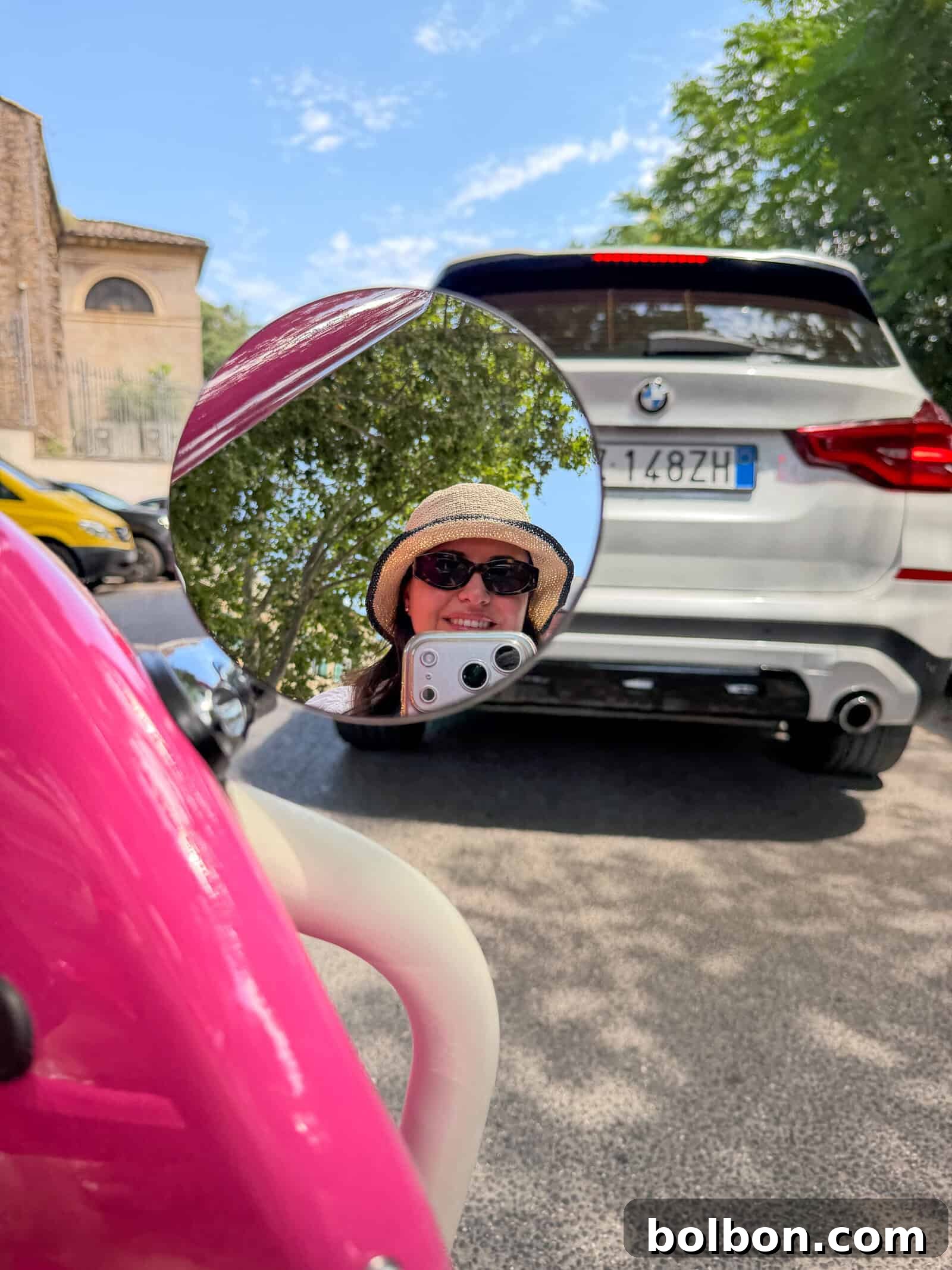 A person wearing sunglasses and a straw hat is reflected in a round mirror attached to a pink scooter. They are taking a photo. In the background, there’s a white car parked on a sunny street with trees and buildings.