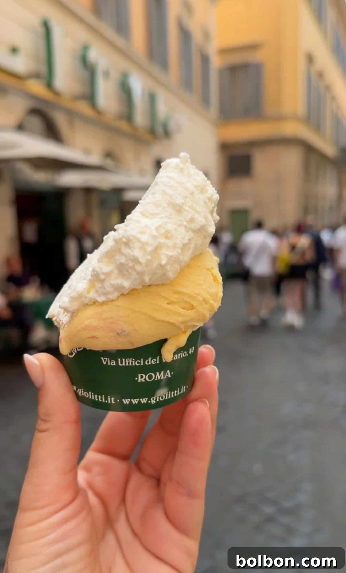 A hand holds a cup of gelato with a large swirl of whipped cream on top; blurred pedestrians and yellow buildings are visible in the background, suggesting a lively street scene in Rome.