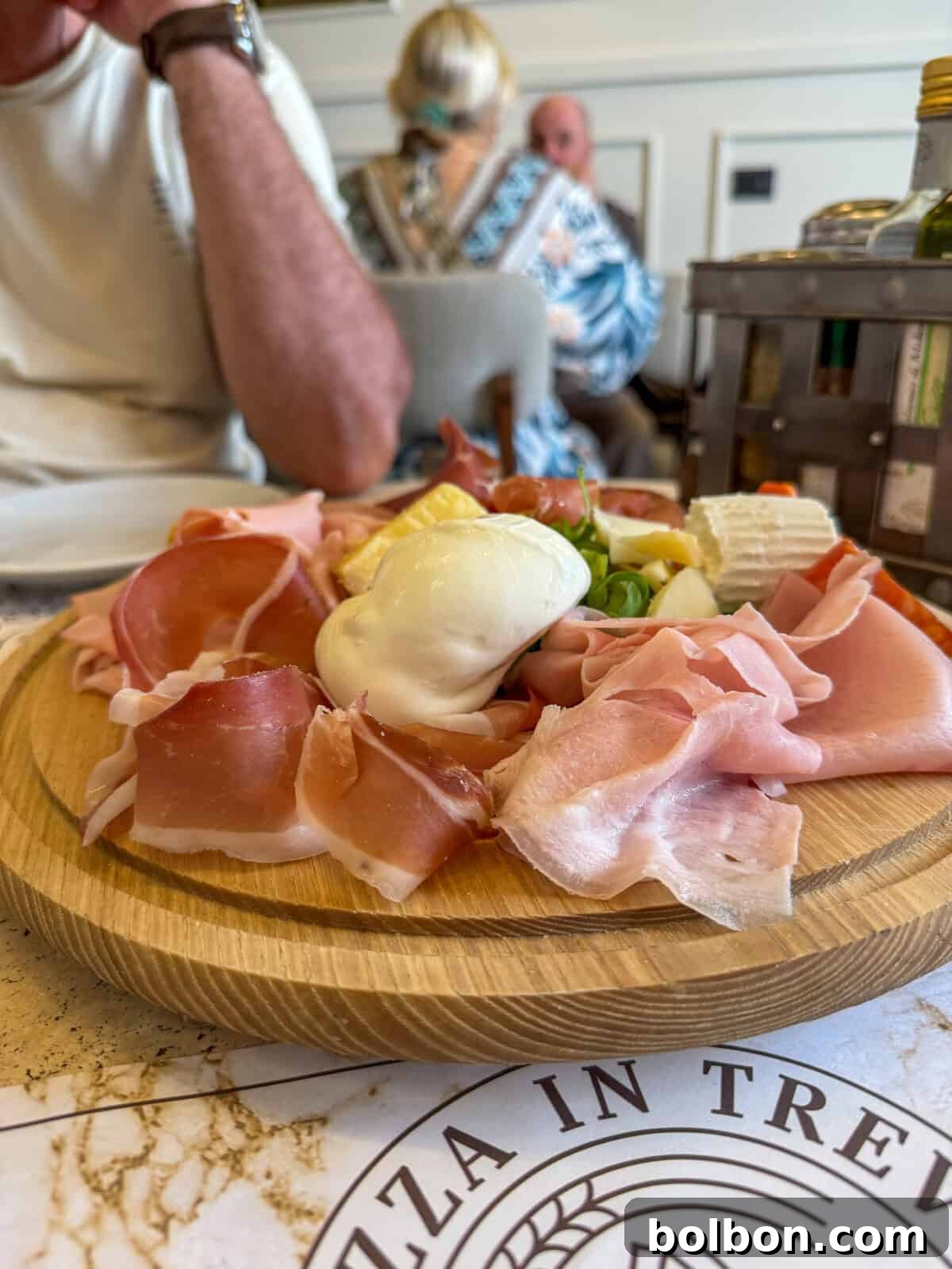 A wooden platter with assorted cured meats, cheese, and a ball of fresh mozzarella is on a table in a restaurant. Diners are seated in the background, and part of a menu is visible in the foreground.