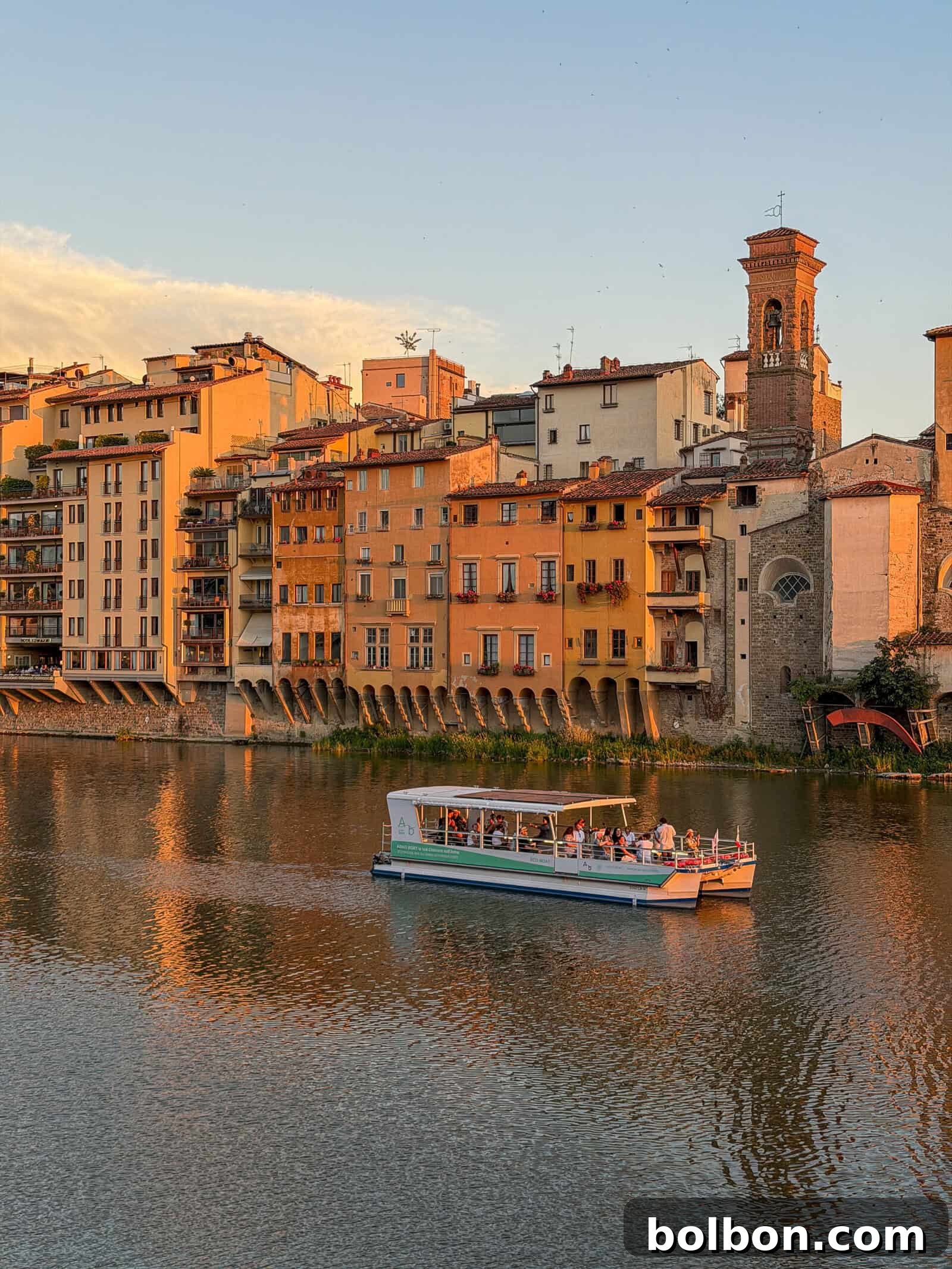 A boat with passengers cruises on a river at sunset, with colorful historic buildings and a tower lining the waterfront in the background.