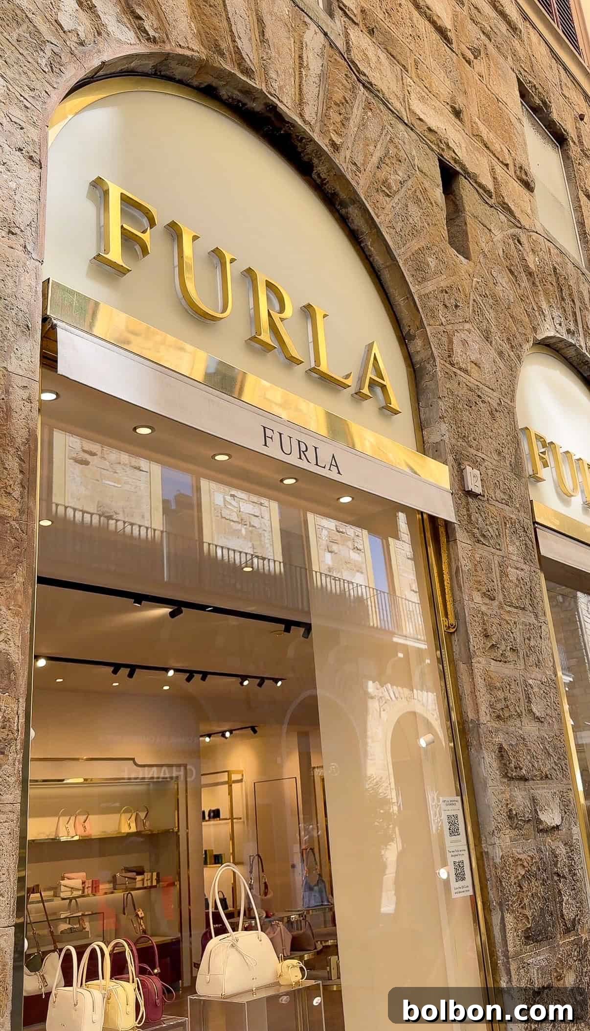 Storefront of a Furla boutique with large gold letters above the entrance, glass windows showing purses and accessories on display, set in a stone building.