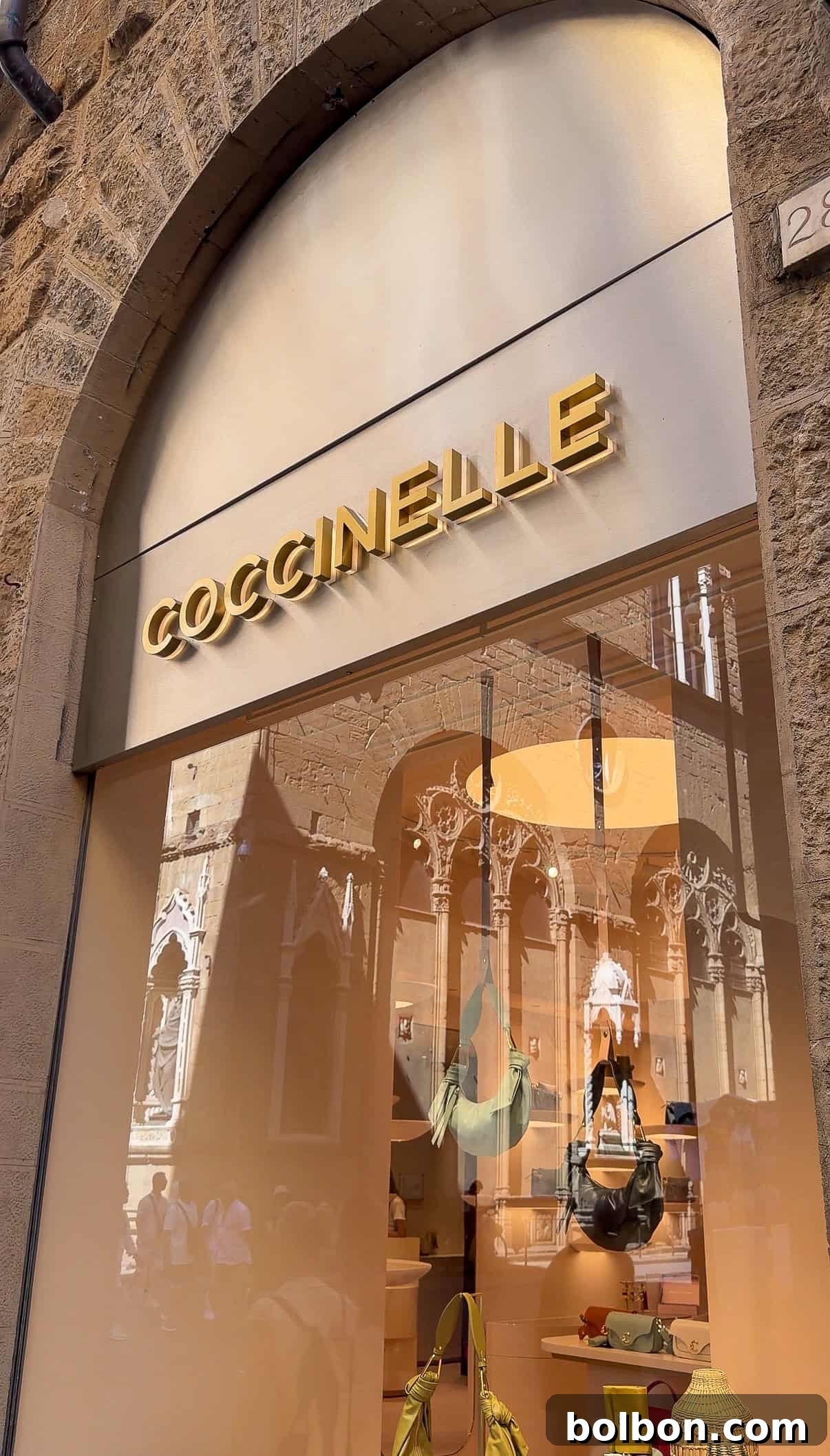 A store window with the sign "COCCINELLE" in gold letters. Handbags and accessories are displayed inside, with reflections of a historic building and people in the glass.