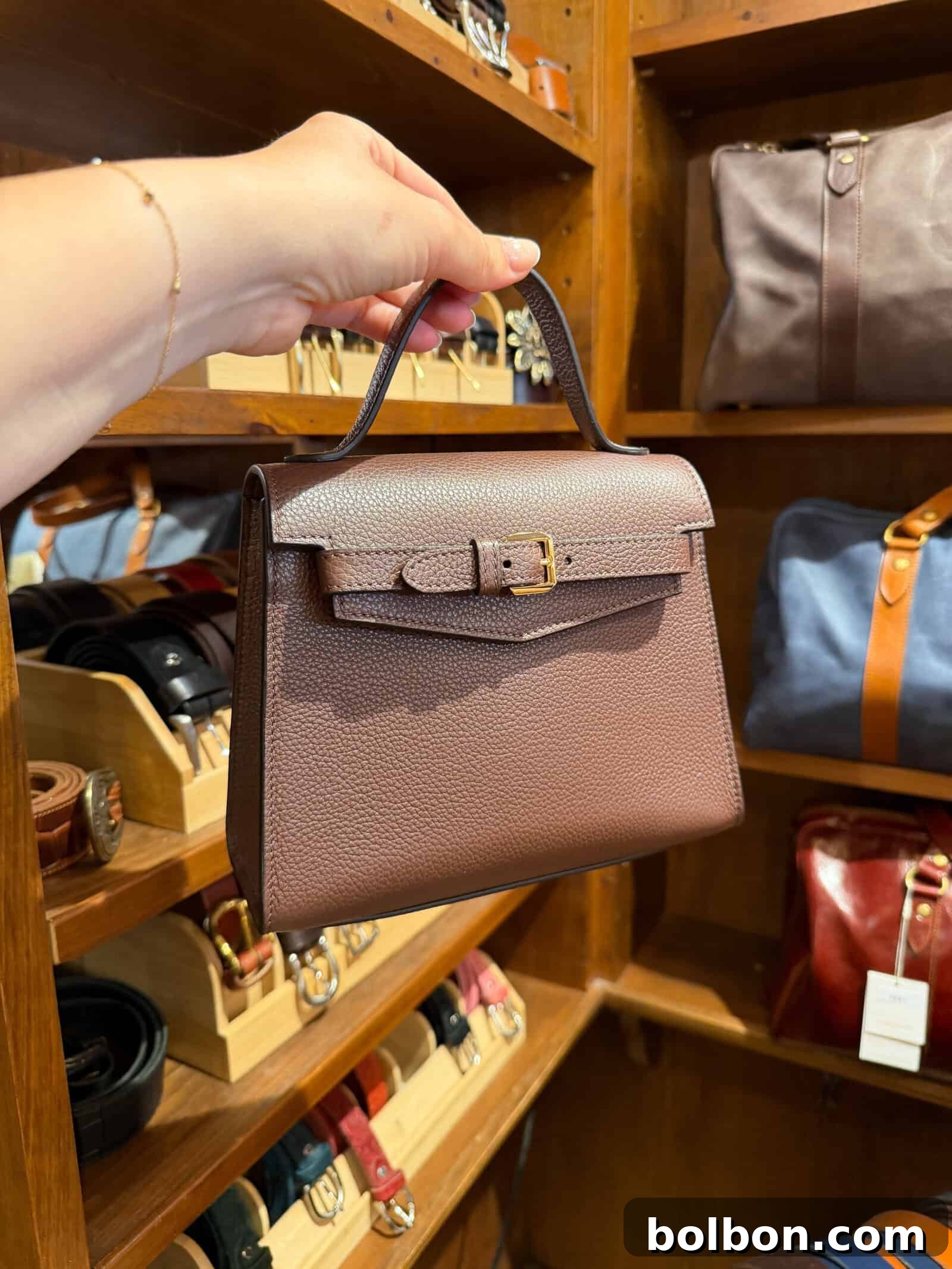 A hand holds a small brown leather handbag with a top handle and buckle, displayed in a store with shelves of belts and bags in the background.