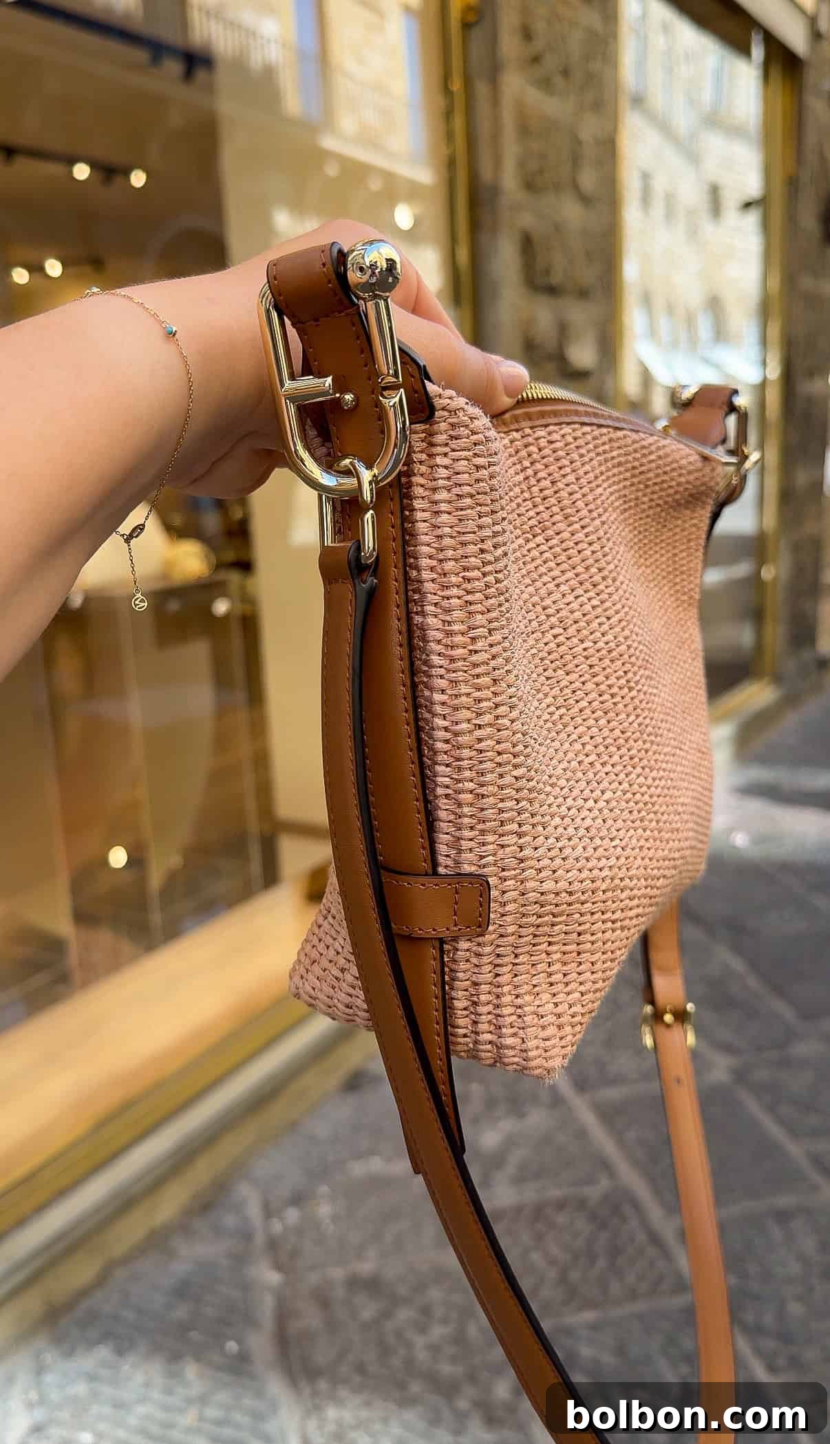A hand holds a woven beige handbag with brown leather straps and gold hardware in front of a shop window on a cobblestone street.