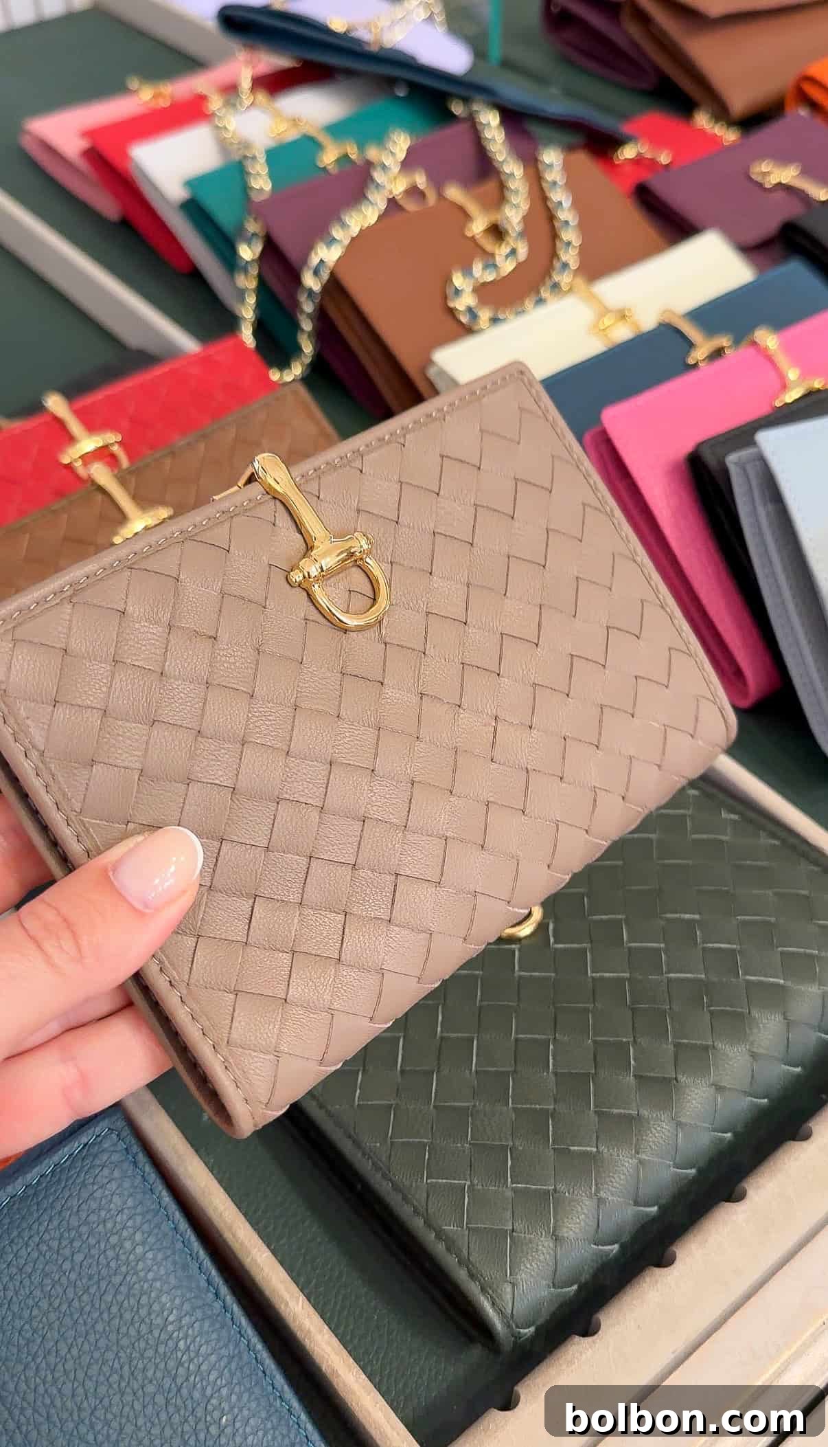 A hand holds a beige woven leather wallet with a gold clasp, displayed among wallets in various colors and styles on a table.