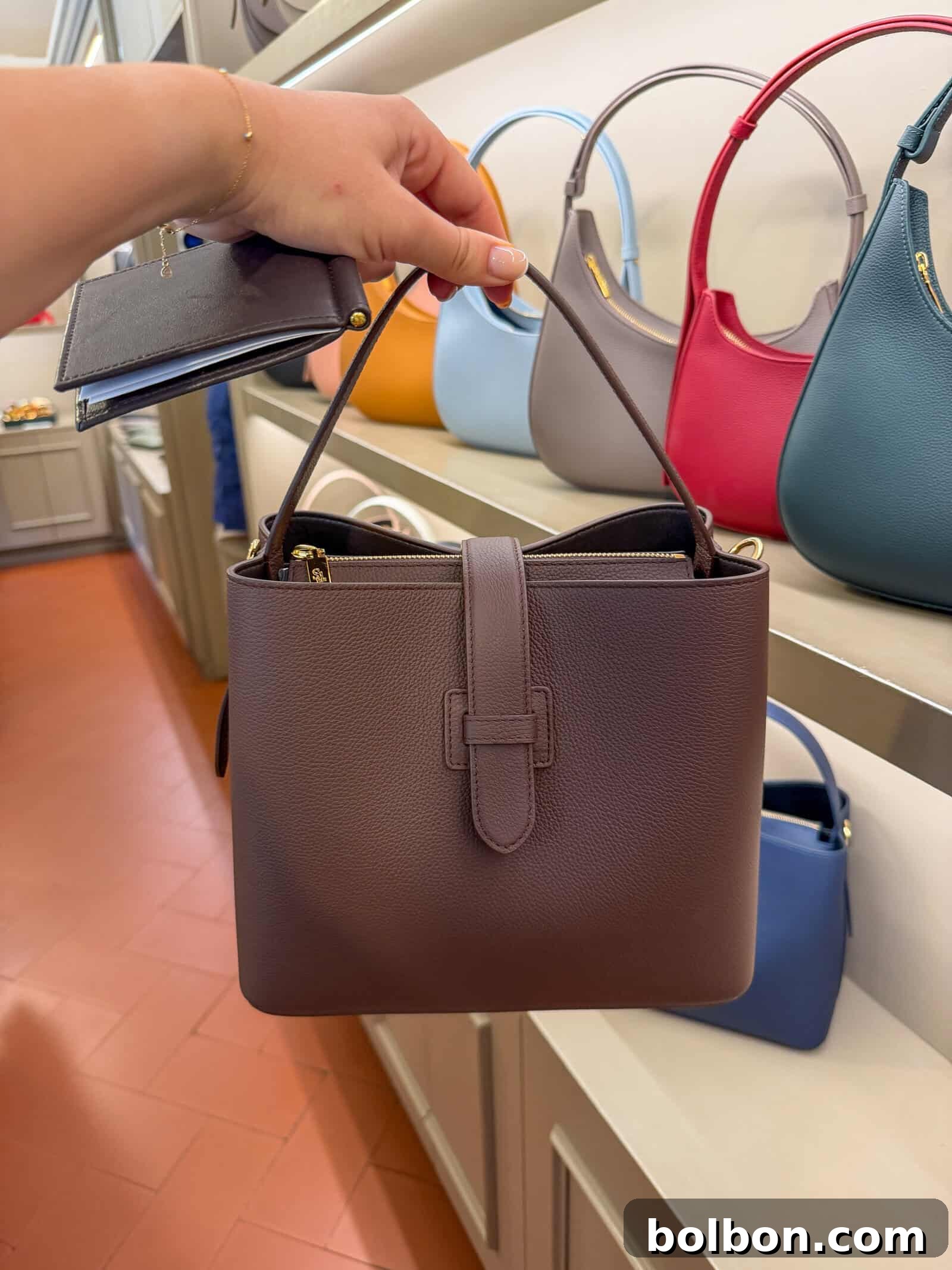 A hand holds a brown leather handbag with a strap in a store. Other colorful handbags are displayed on shelves in the background.