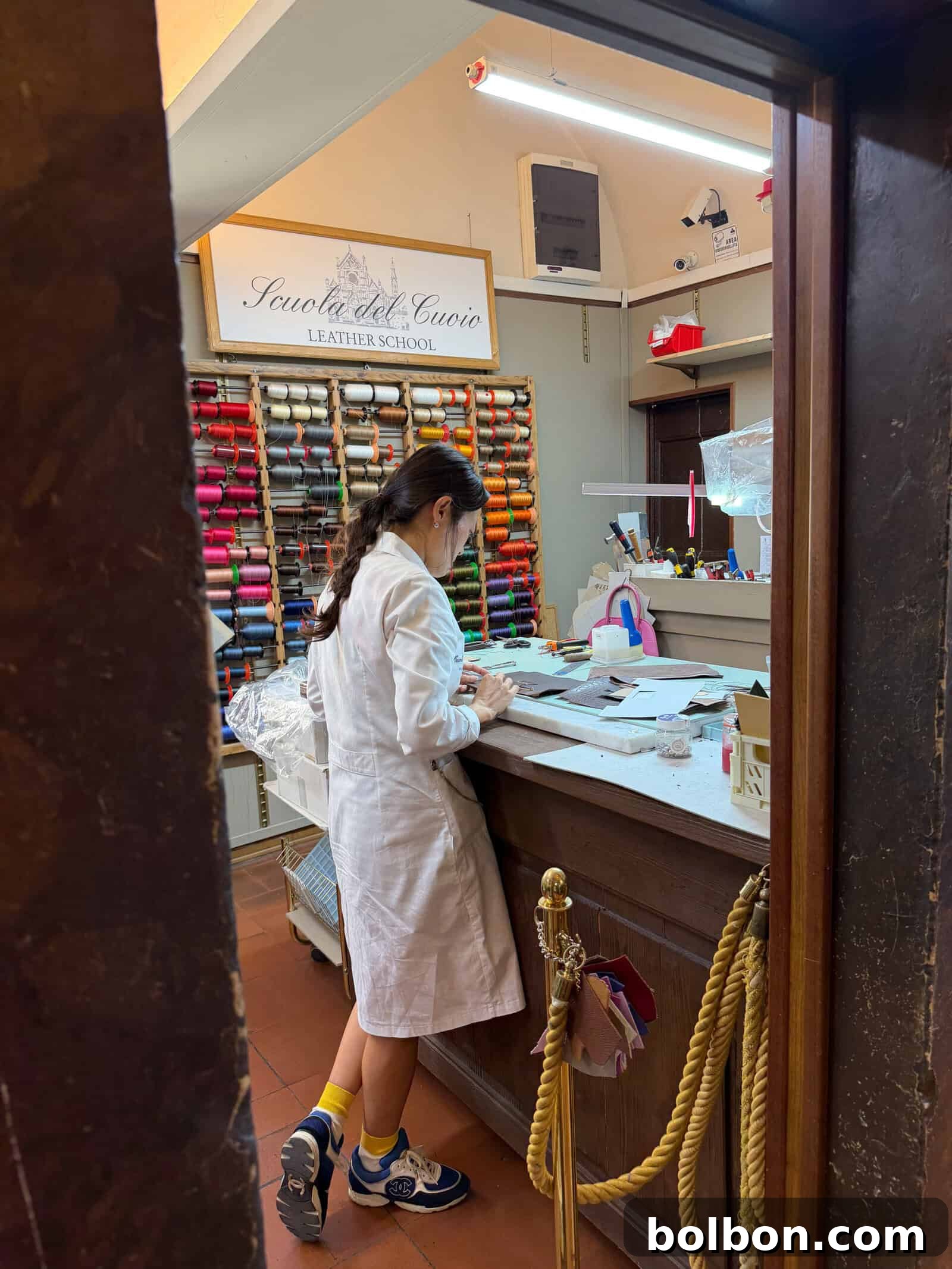 A person in a white lab coat works at a counter in a leather school, with colorful threads organized on the wall behind them and various leatherworking tools on the desk.