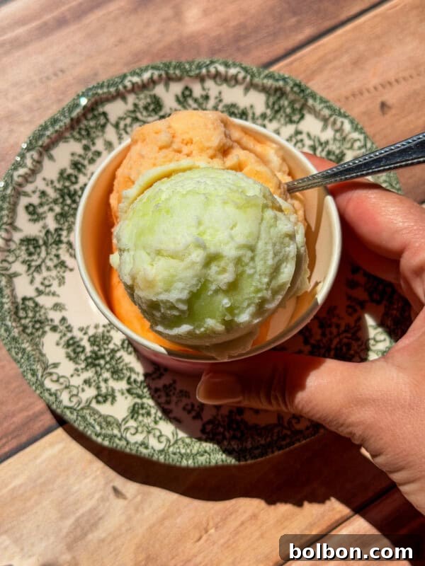 A hand holds a spoon in a bowl with two scoops of ice cream—one green and one orange.