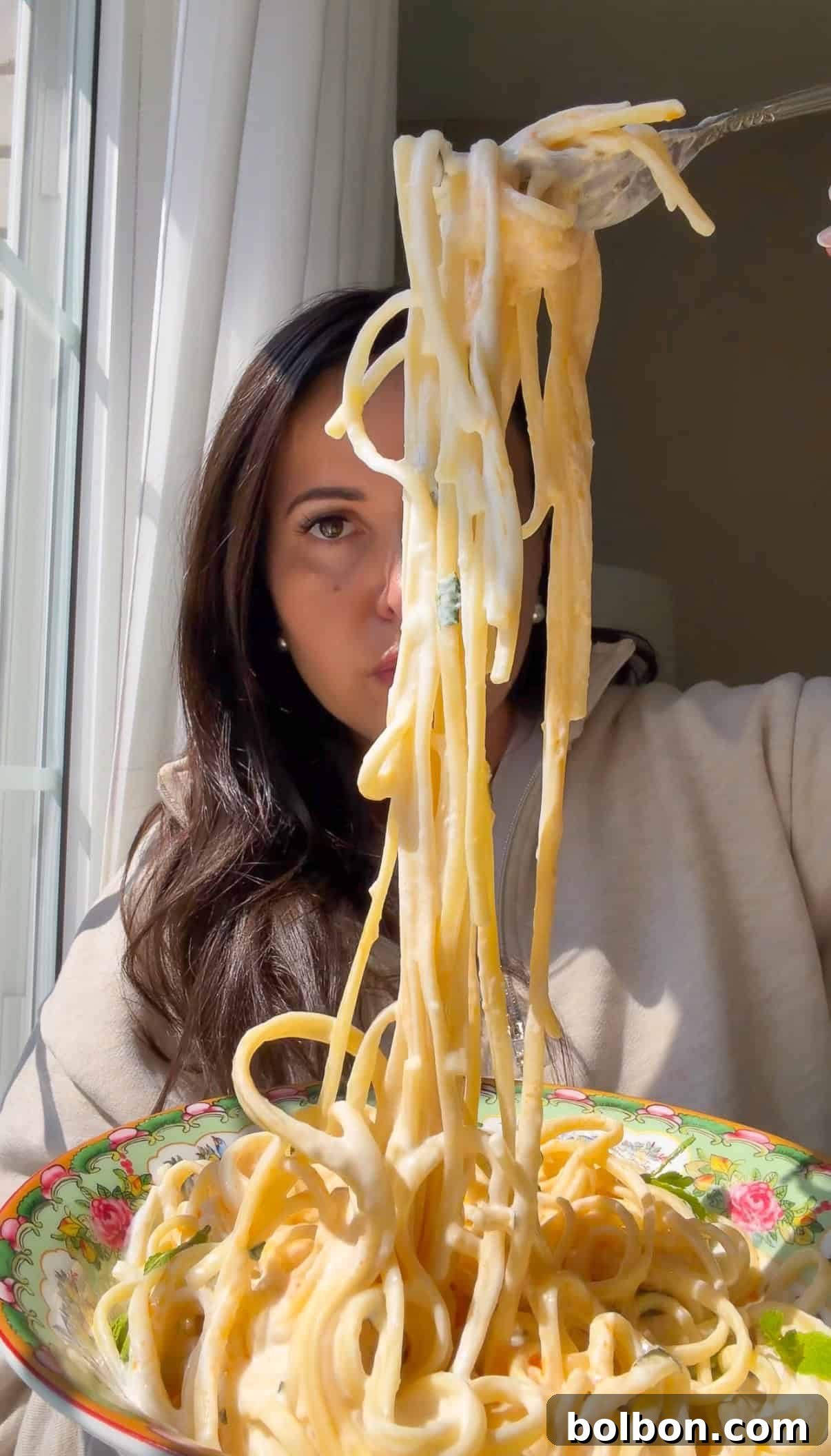 TikTok Viral Yogurt Noodles Recipe 2 A woman in a beige top lifts a forkful of creamy yogurt noodles from a colorful floral bowl, with sunlight streaming through a window beside her.