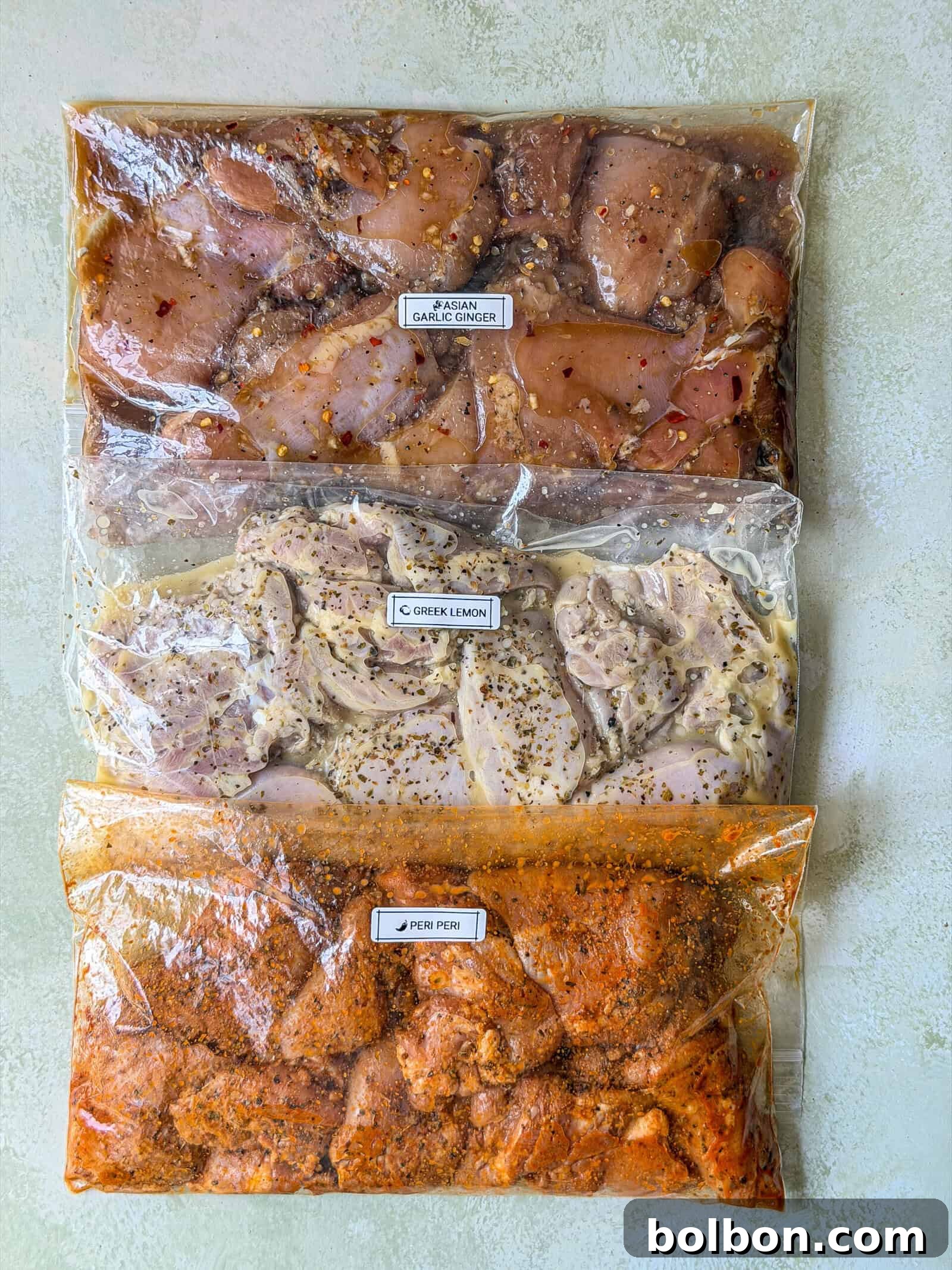 3 Chicken Marinades for Meal Prep and Freezer Cooking 2 Three ziplock bags filled with marinated raw chicken breast, labeled Asian Garlic Ginger, Greek Lemon, and Peri Peri, are placed on a light-colored surface—showcasing a delicious variety of chicken marinades ready for any meal.