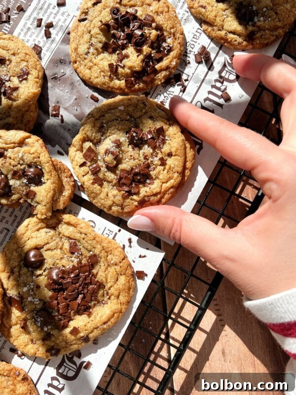 A hand reaches for a Brown Butter Chocolate Chip Cookie on a cooling rack lined with newspaper-style parchment, surrounded by other cookies crowned with chocolate chunks as sunlight casts inviting shadows across the scene.