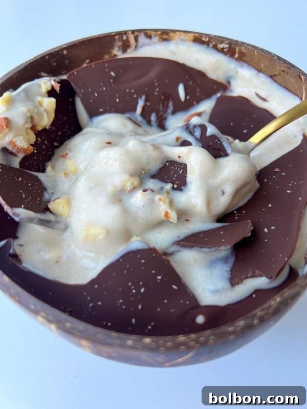 A bowl filled with creamy banana ice cream, topped with large pieces of dark chocolate and sprinkled with chopped nuts, creates a Magic Shell Smoothie Bowl, served with a gold spoon resting inside.