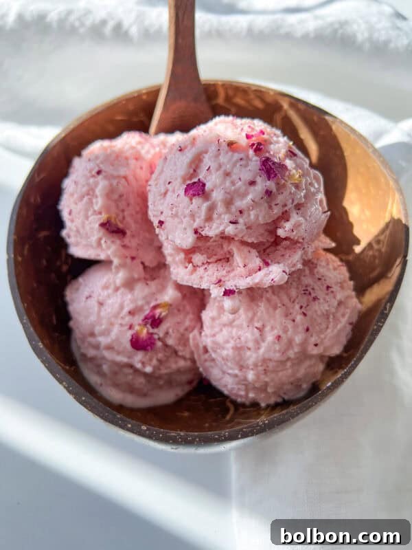 A coconut bowl filled with four scoops of light pink Cottage Cheese Ice Cream, garnished with small edible flower petals, on a white surface with sunlight casting soft shadows.