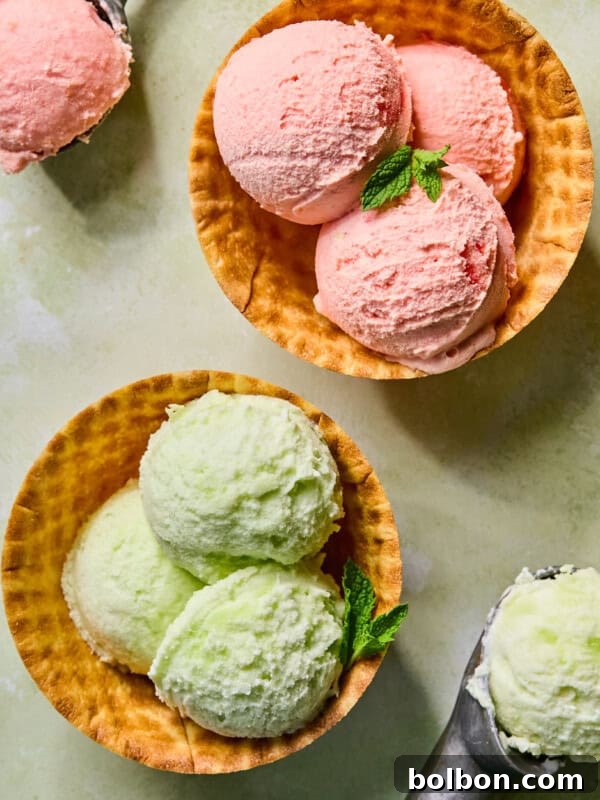 Two waffle bowls filled with three scoops of ice cream each, one with pink ice cream and the other with light green, both garnished with fresh mint. Metal ice cream scoops and extra ice cream are nearby.