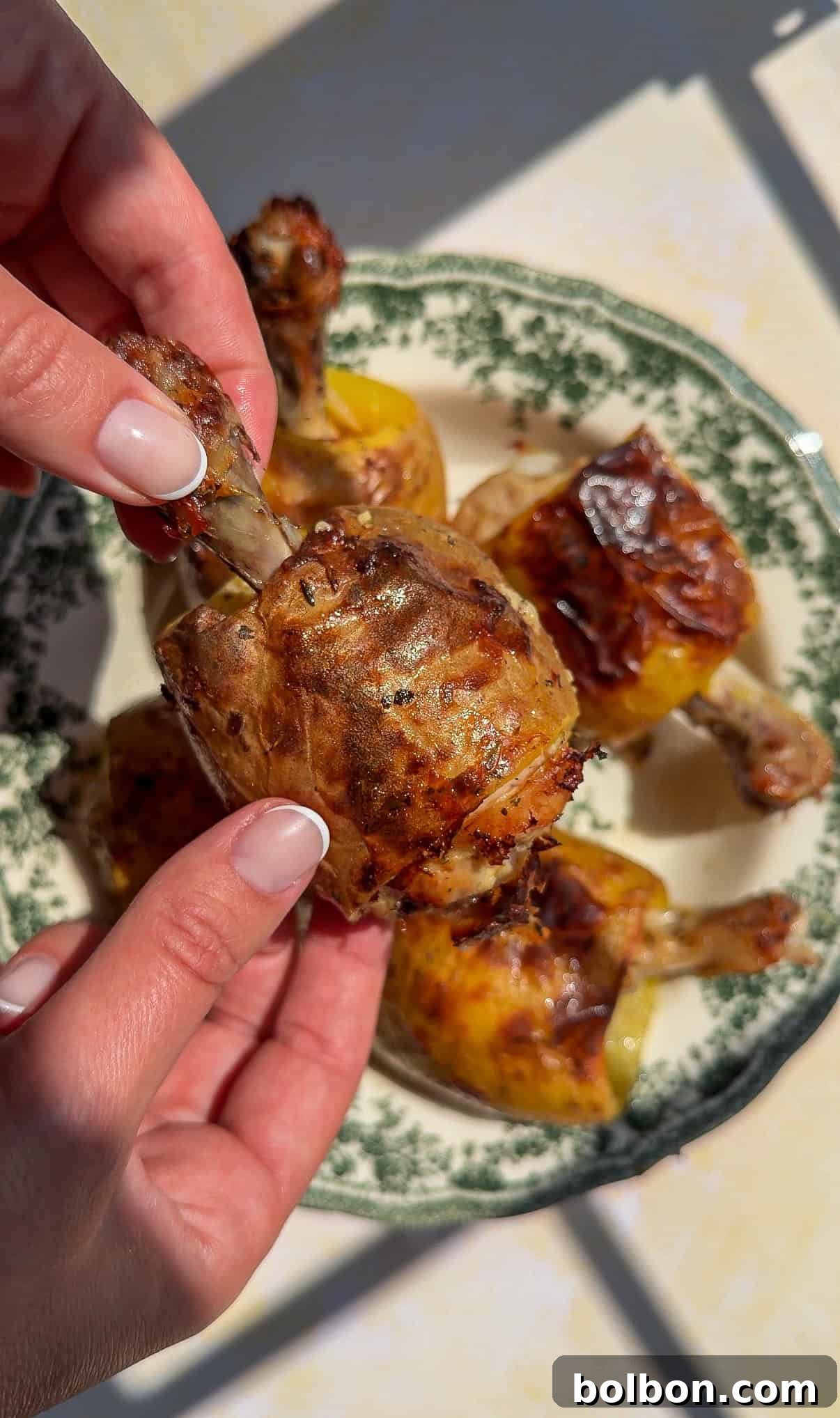 Two hands holding a cooked chicken drumstick with crispy skin over a green and white patterned plate with more drumsticks on it