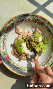 A hand holds a fork over a colorful floral plate containing tuna, avocado, ricotta cheese, and pesto, with sunlight casting shadows across the scene.