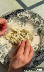 Two hands fold a rice paper sheet filled with shredded cheese and herbs on a decorative plate with a black floral pattern.