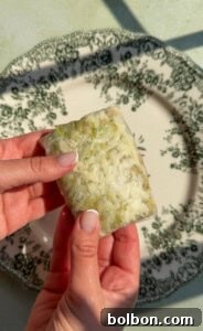 Two hands hold a rectangular block of rice and green vegetables wrapped in translucent paper, over a decorative white plate with a dark floral pattern.