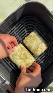Two hands place two rectangular pieces of food topped with shredded cheese, wrapped in plastic, into an air fryer basket.